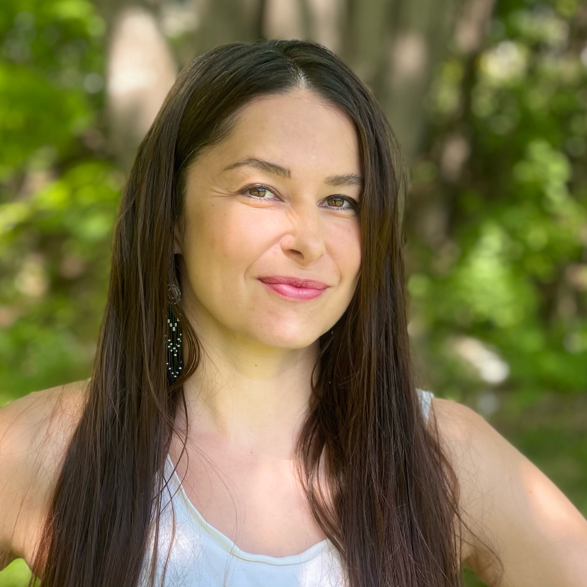 A metis woman standing outside with green leaves behind her. She is wearing a blue tank top and her long brown hair is down. She has a small smile and pink lips.