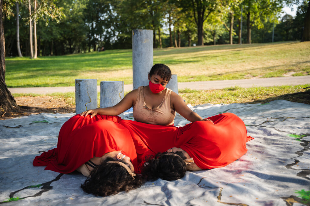 Woman craddling two people under a red cloth. She sits on a large tarp painted like concrete, with cracks of grass and gold poking through