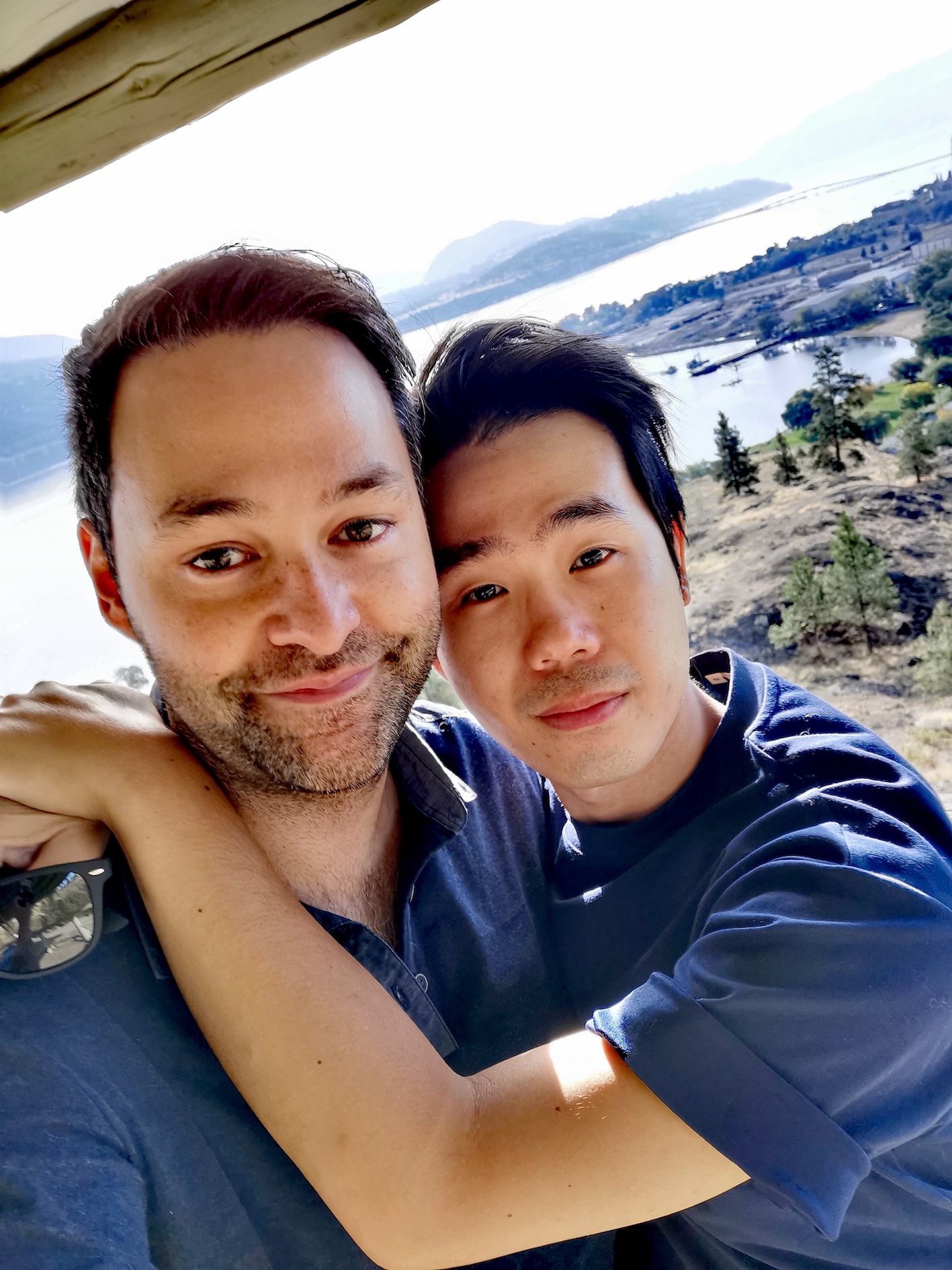 Two men closed mouth smile at the camera. Jeff Ho has his arms casually drapped around his partner Pierre