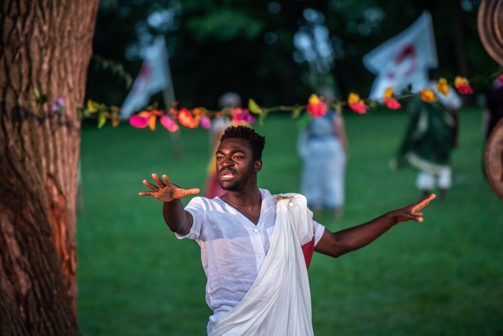 Production still from Portia's Julius Cesar - Kwaku a young black man is dressed in a white toga. He is reaching passionately into the distance