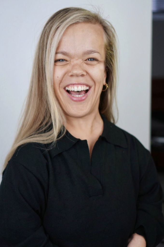 ALT: A headshot of Alexia, a white, Disabled woman with blonde hair. She is mid-laugh in the photo. She's wearing a black shirt with gold hoop earrings. The background is white. Photograph by Lindsay Wu.